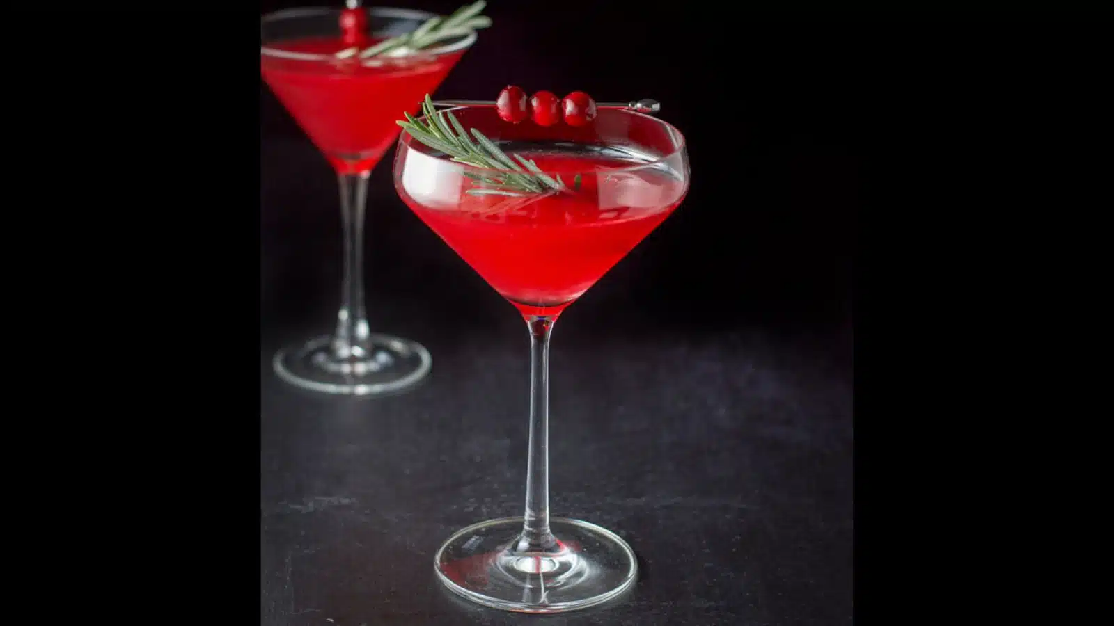 Two martini glasses filled with the red holiday cocktail with rosemary and cranberries as garnish