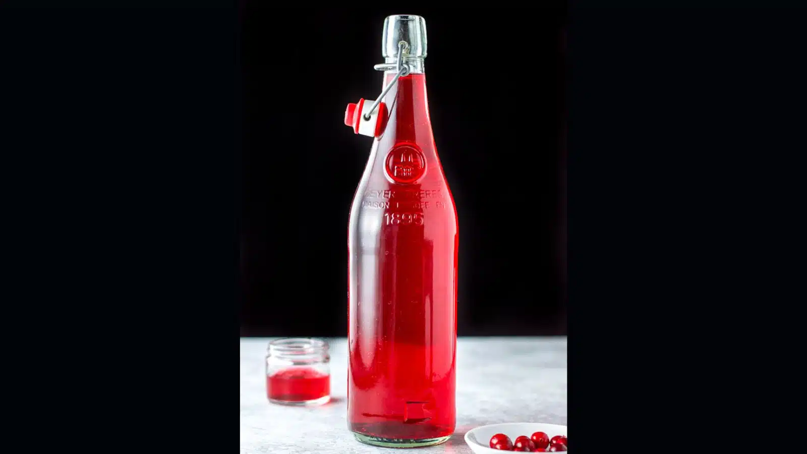 A large glass bottle filled with cranberry vodka along with a little in a jar and some cranberries in a white bowl