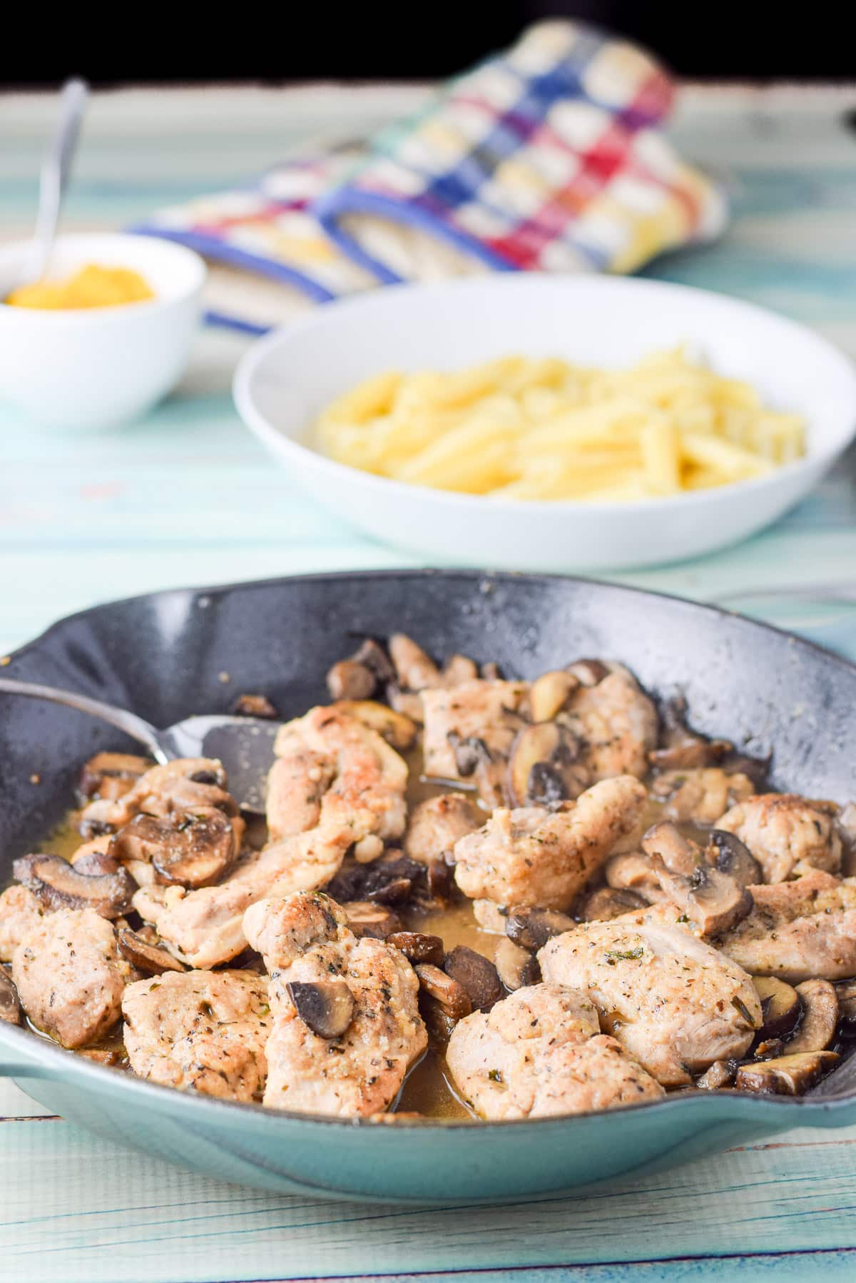 Pasta in a bowl behind a green saute pan filled with a saucy chicken with mushrooms