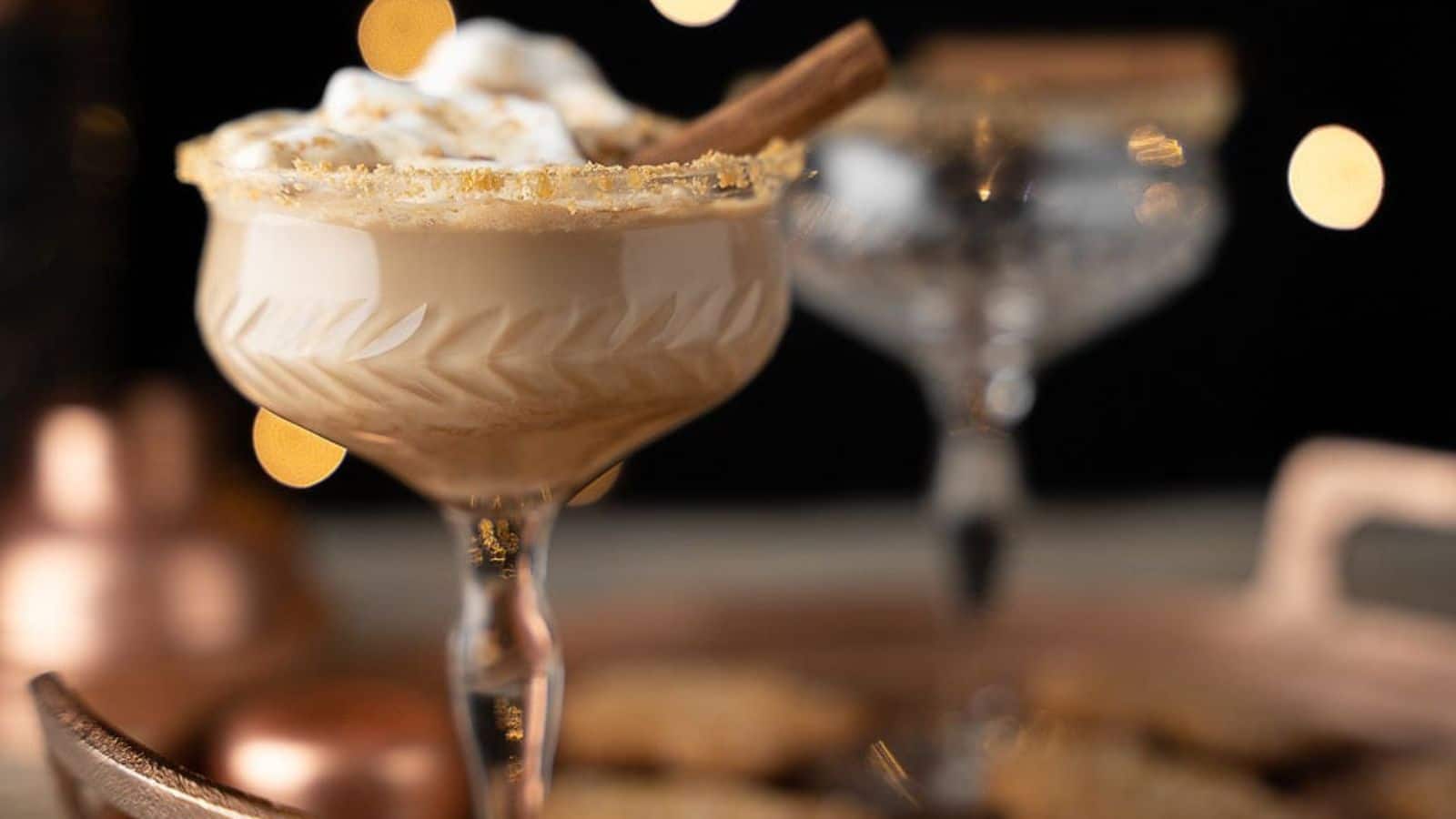Two coupe glasses one filled with the chocolate cocktail with gingerbread crumbs on the rim, whipped cream and a cinnamon stick