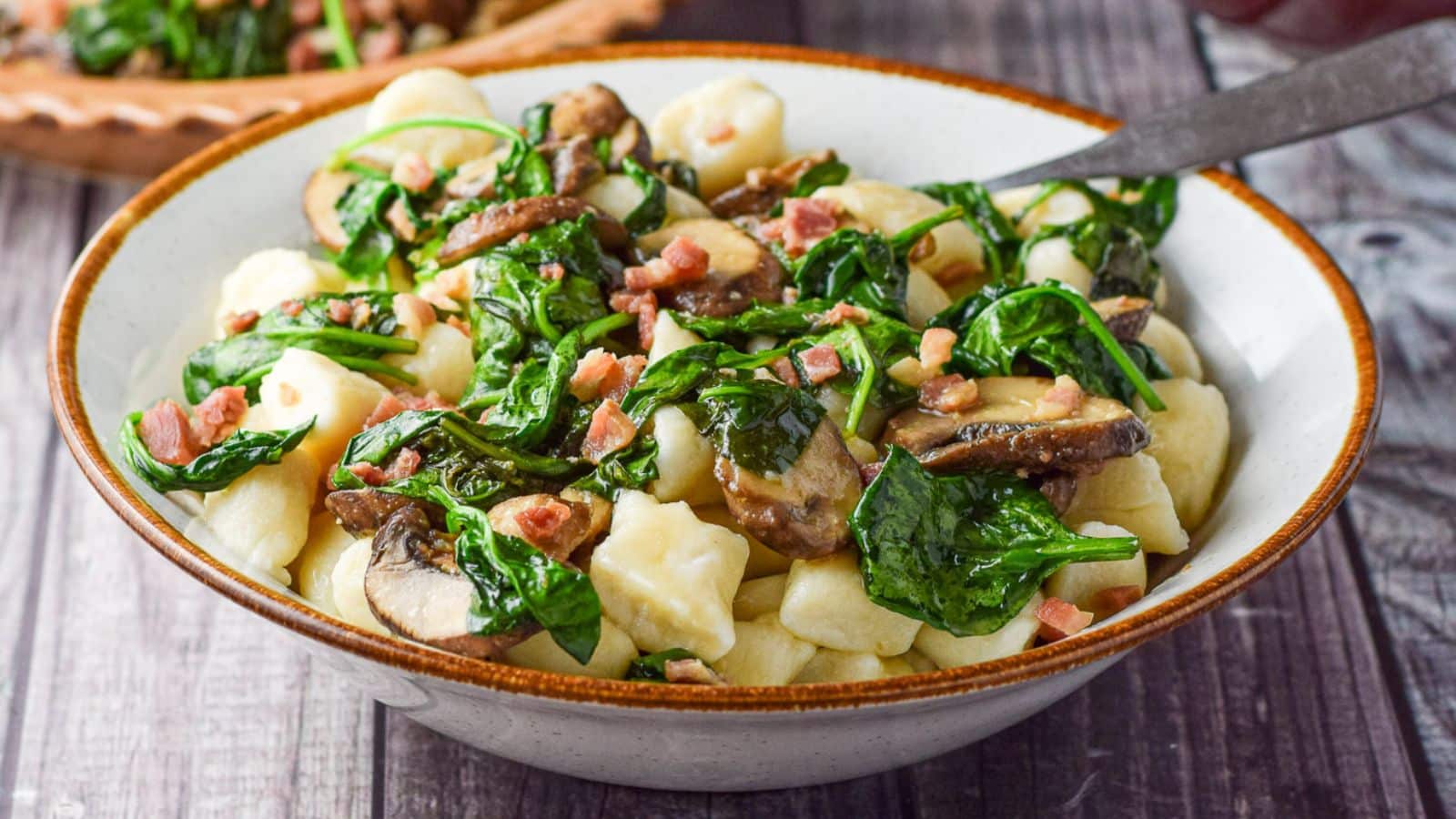 A brown rimmed bowl with gnocchi, spinach and pancetta and a spoon