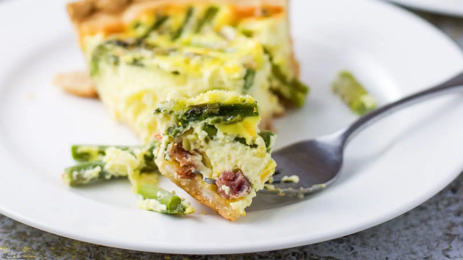 a white small plate with a piece of quiche on it with a fork with a piece showing asparagus and ham