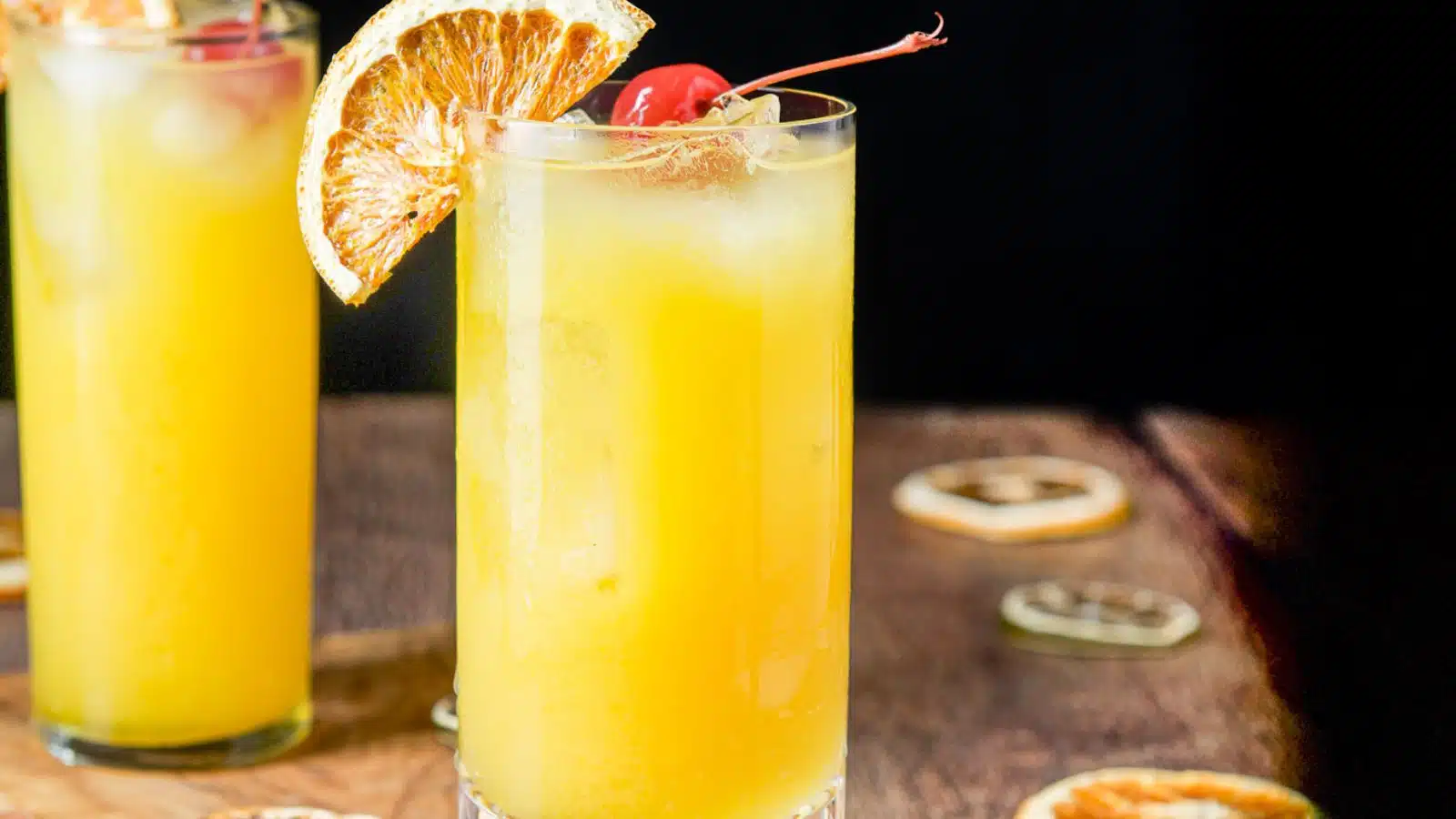 two tall, thin glasses with an orange juice cocktail in it with an orange wheel and cherry as garnish