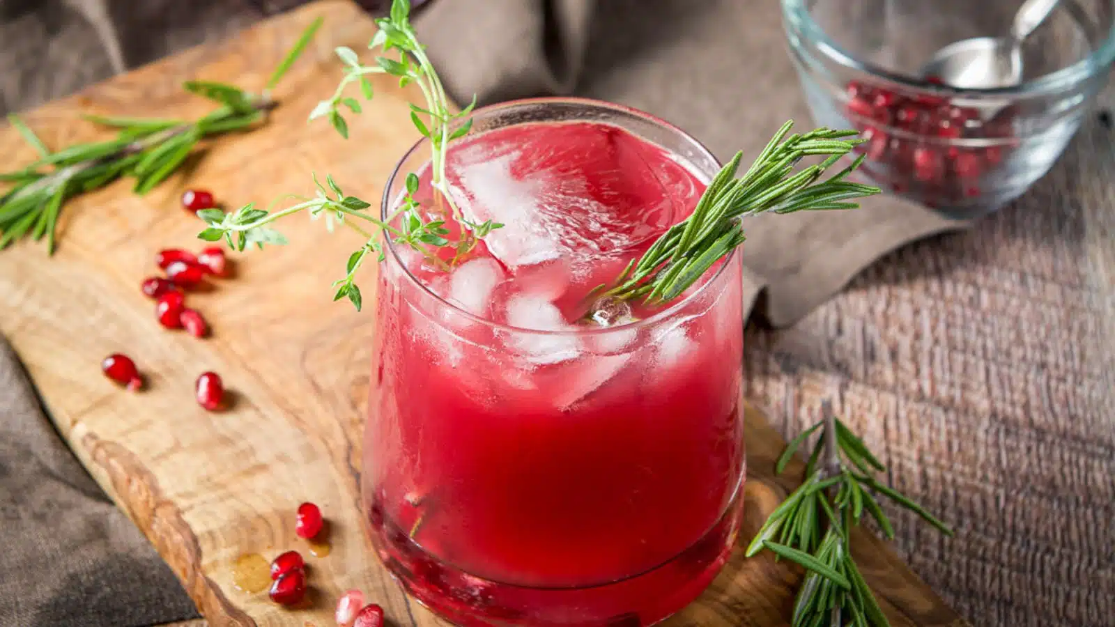 a board with rosemary sprigs and pomegranate seeds on it along with the red cocktail