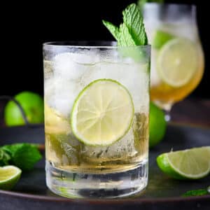 a square photo of a lime cocktail with a lime wheel and mint as garnish. It's on a metal tray with lime wedges and more mint