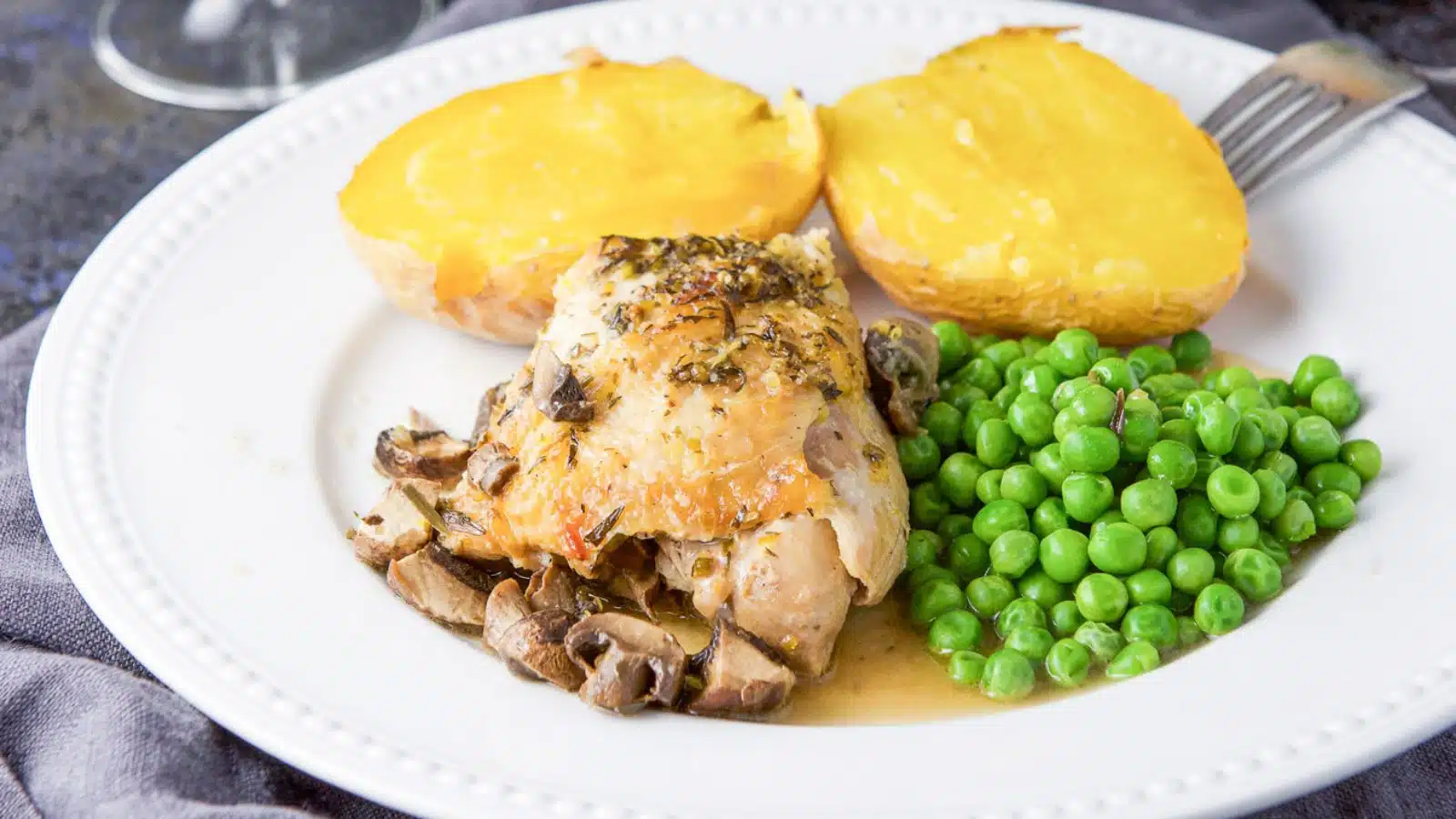 A white beaded plate with a chicken thigh with mushrooms and sauce, peas and baked potato