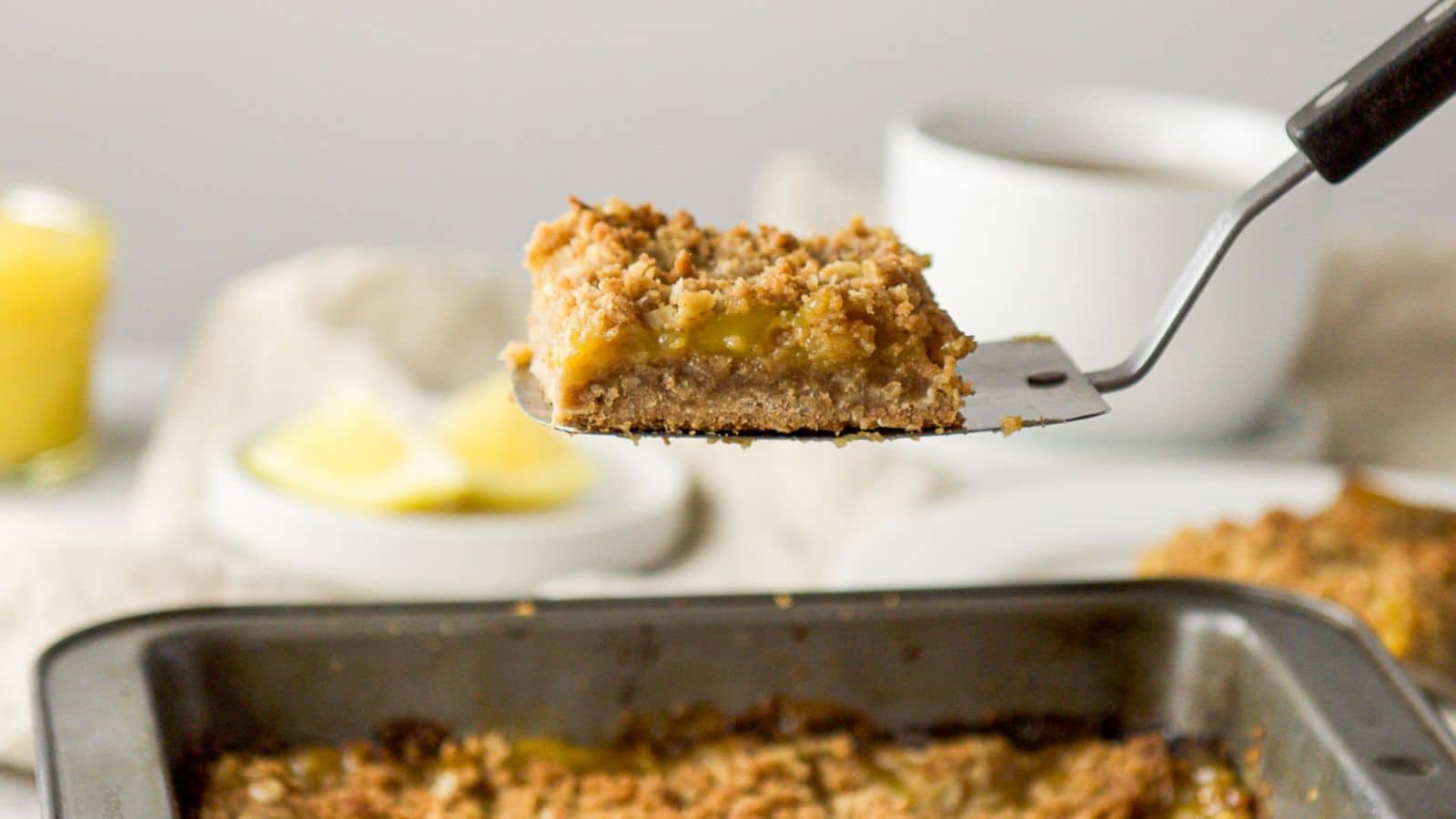 a spatula in the air with a lemon bar on it held over the pan with coffee and lemon in the back