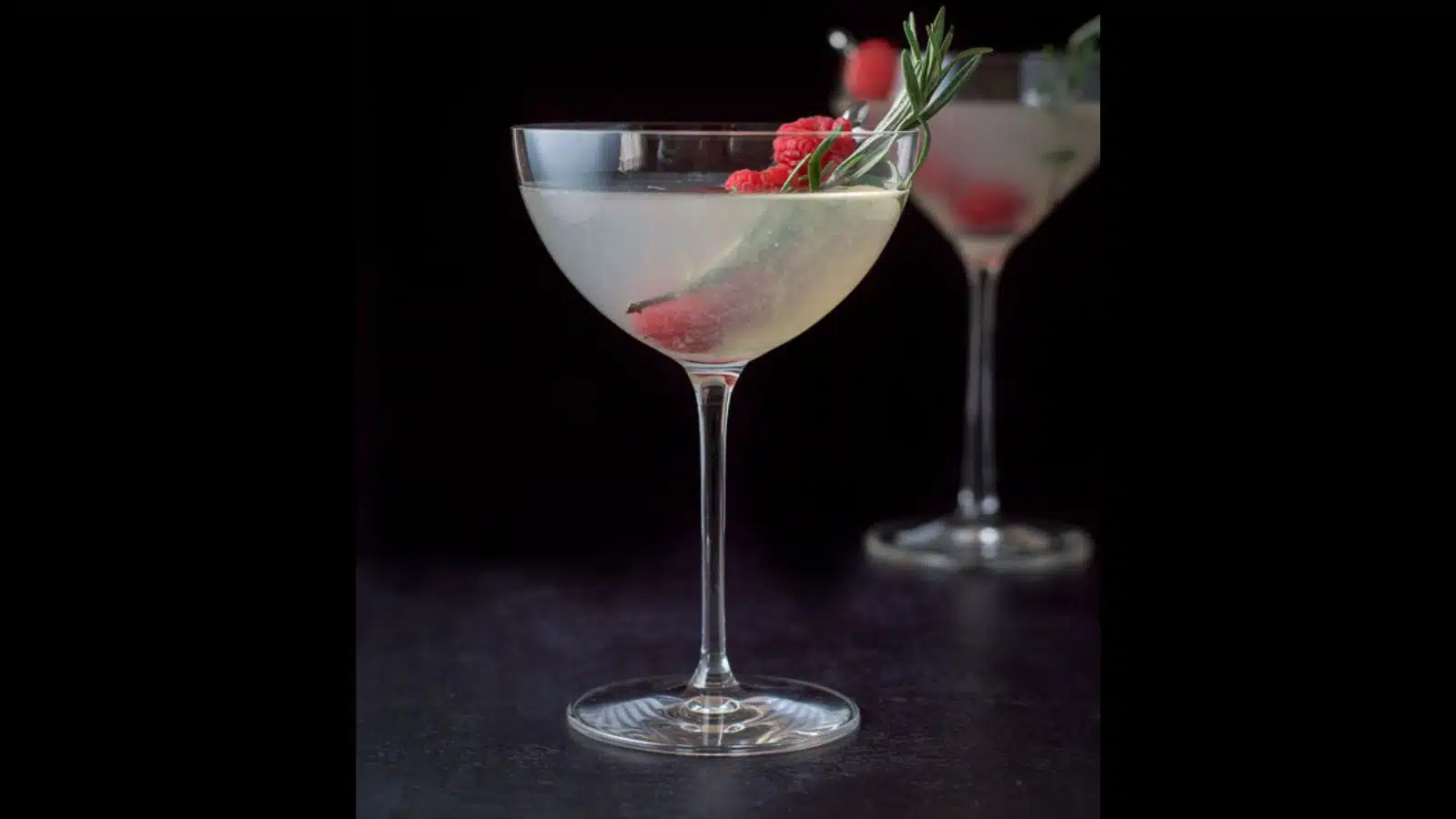 Two martini glasses filled with the festive cocktail with rosemary sprigs and raspberries