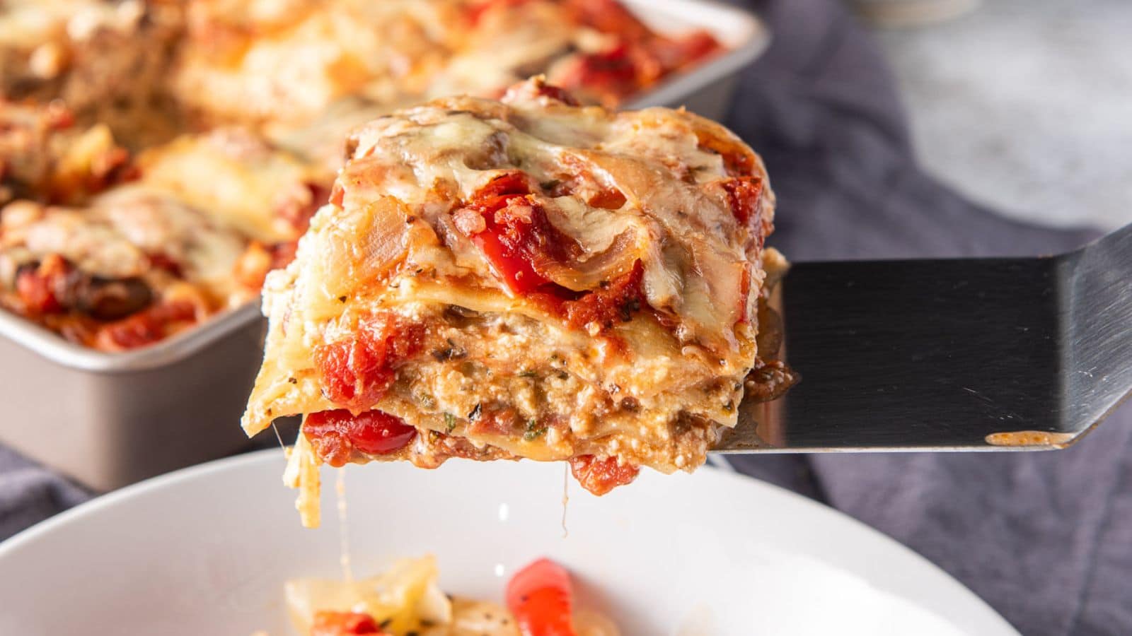 A spatula with a piece of lasagna on it held over a plate with the pan in the back