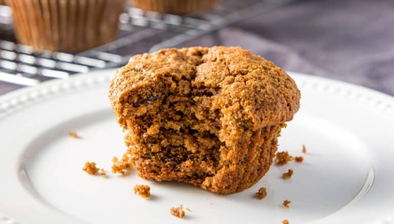 A white plate with a muffin on it with a bite taken out of it along with crumbs and a wire rack in the back with more muffins on it