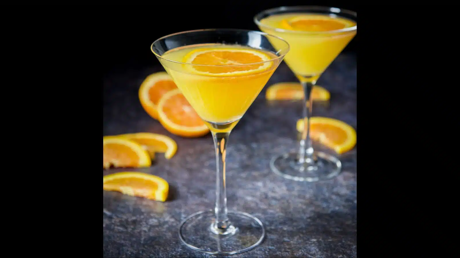 Two glasses of an orange drink with orange wheels in it for garnish and more oranges on the table