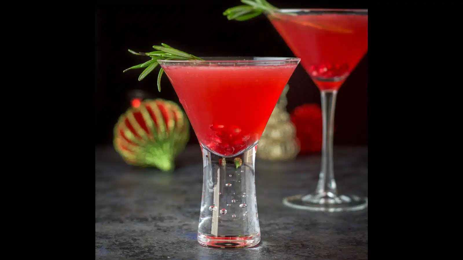 Two martini glasses filled with pomegranate seeds, rosemary sprigs in a red drink