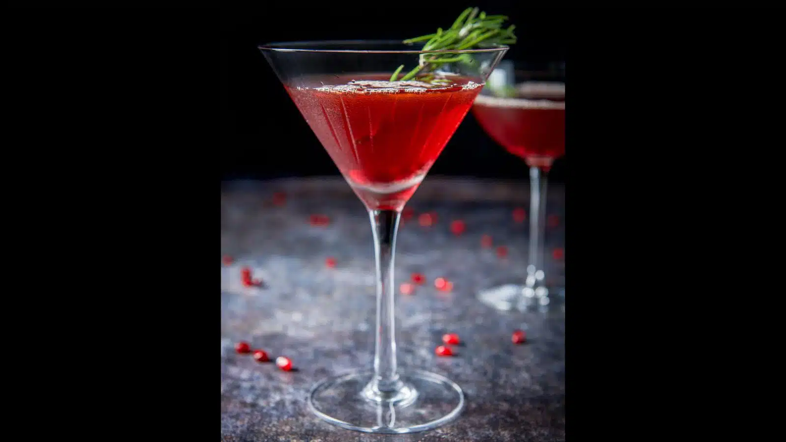 A beveled martini glass filled with the red cocktail with pomegranate seeds on the table and rosemary in the glass