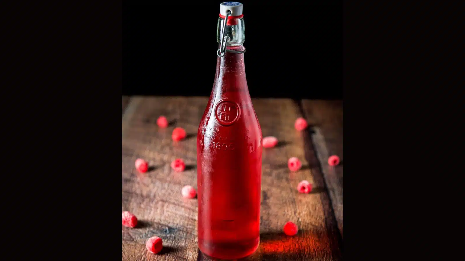 A tall glass bottle with raspberry vodka in it with raspberries on the wood table