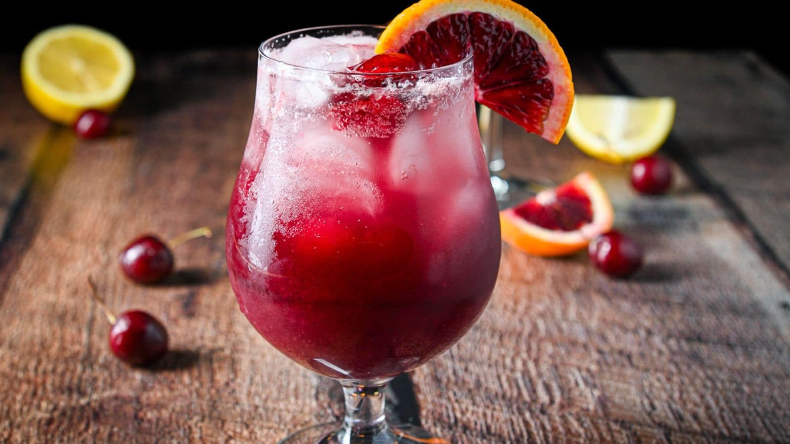 A tulip glass filled with the red wine drink with fruit in the glass along with more fruit on the wooden table