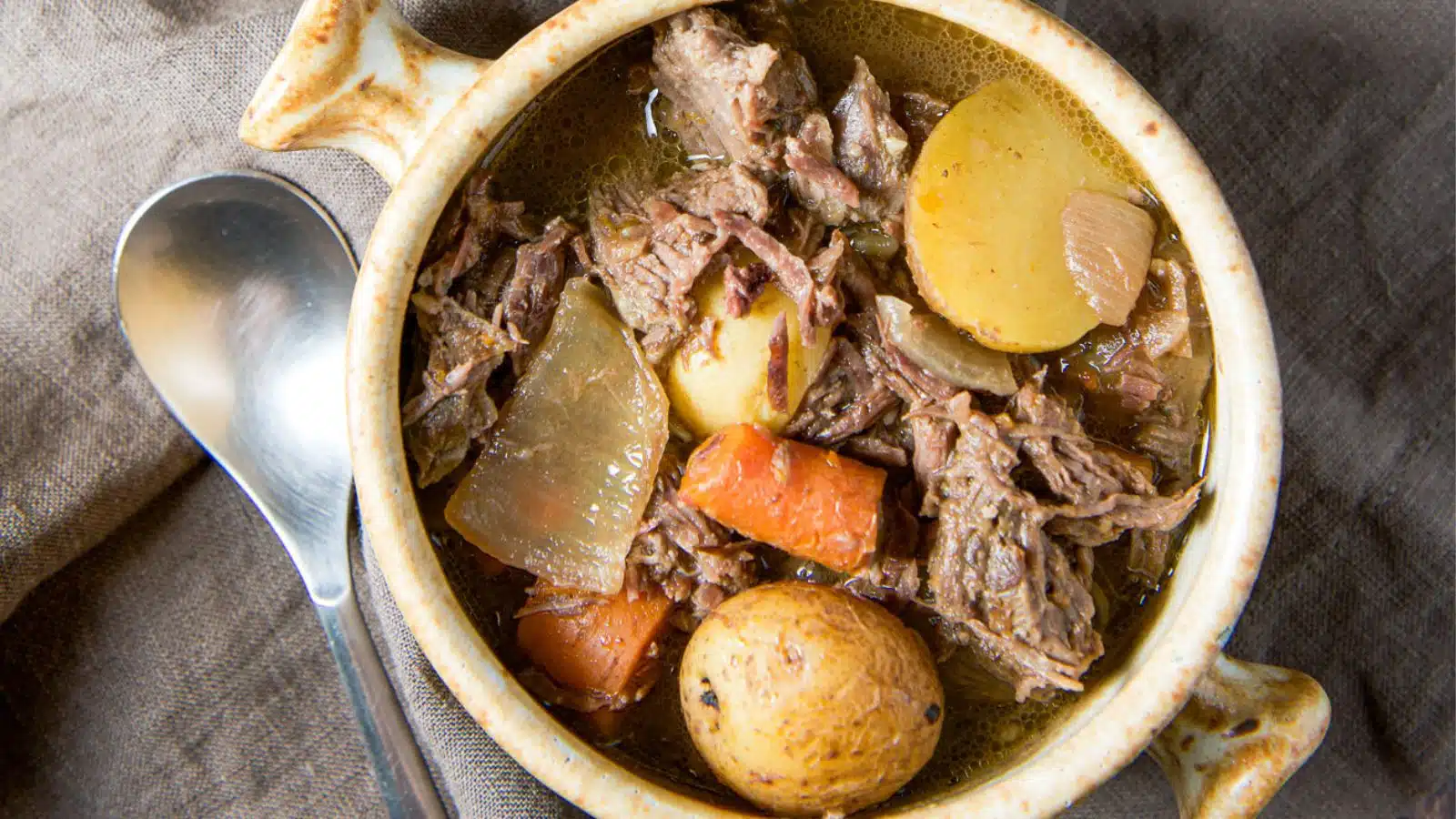 an overhead view of potatoes, carrots and beef pot roast in a bowl with a spoon