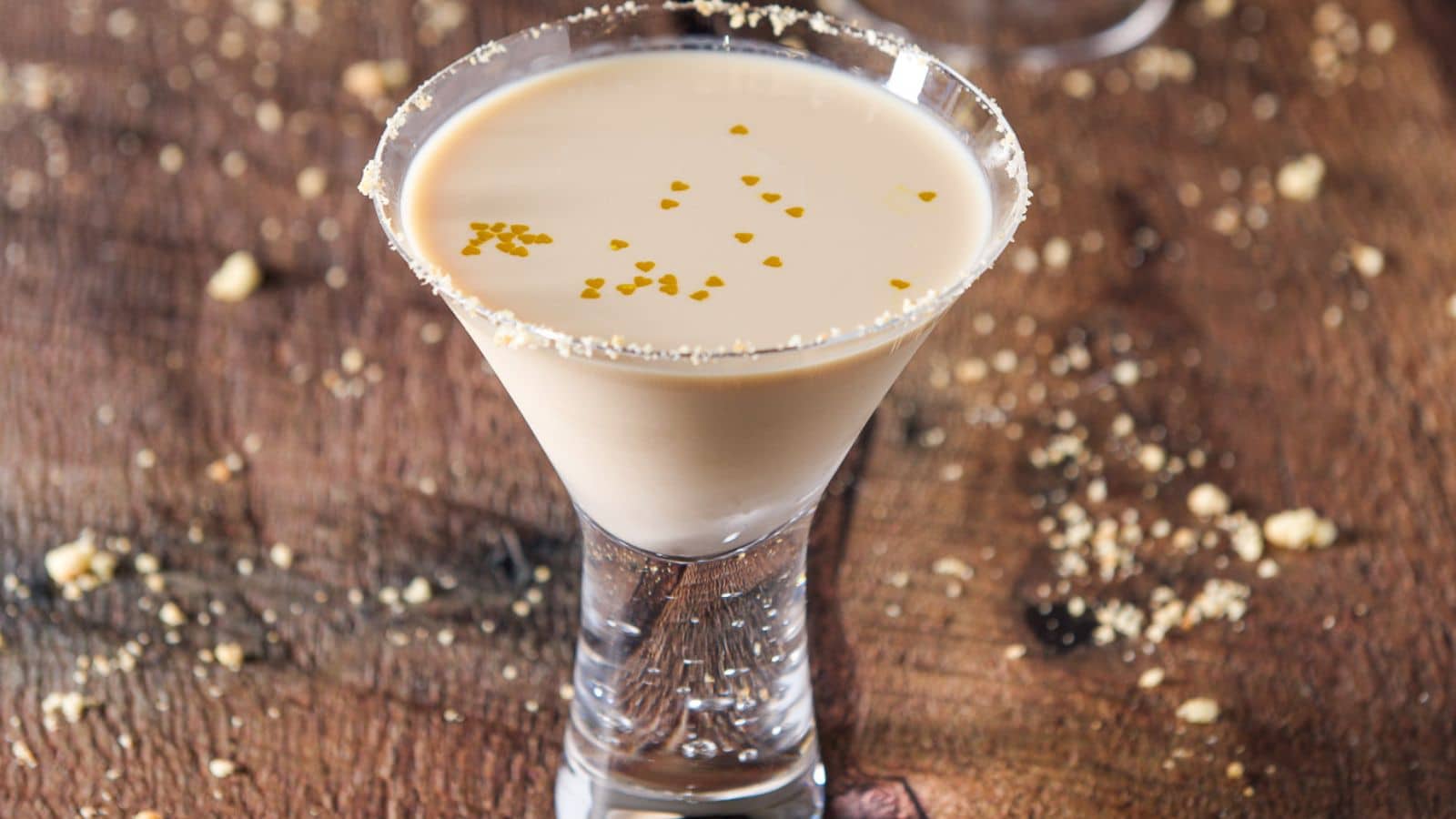 A martini glass with a cream cocktail in it with cookie crumbs on the rim and table and little gold hearts in it