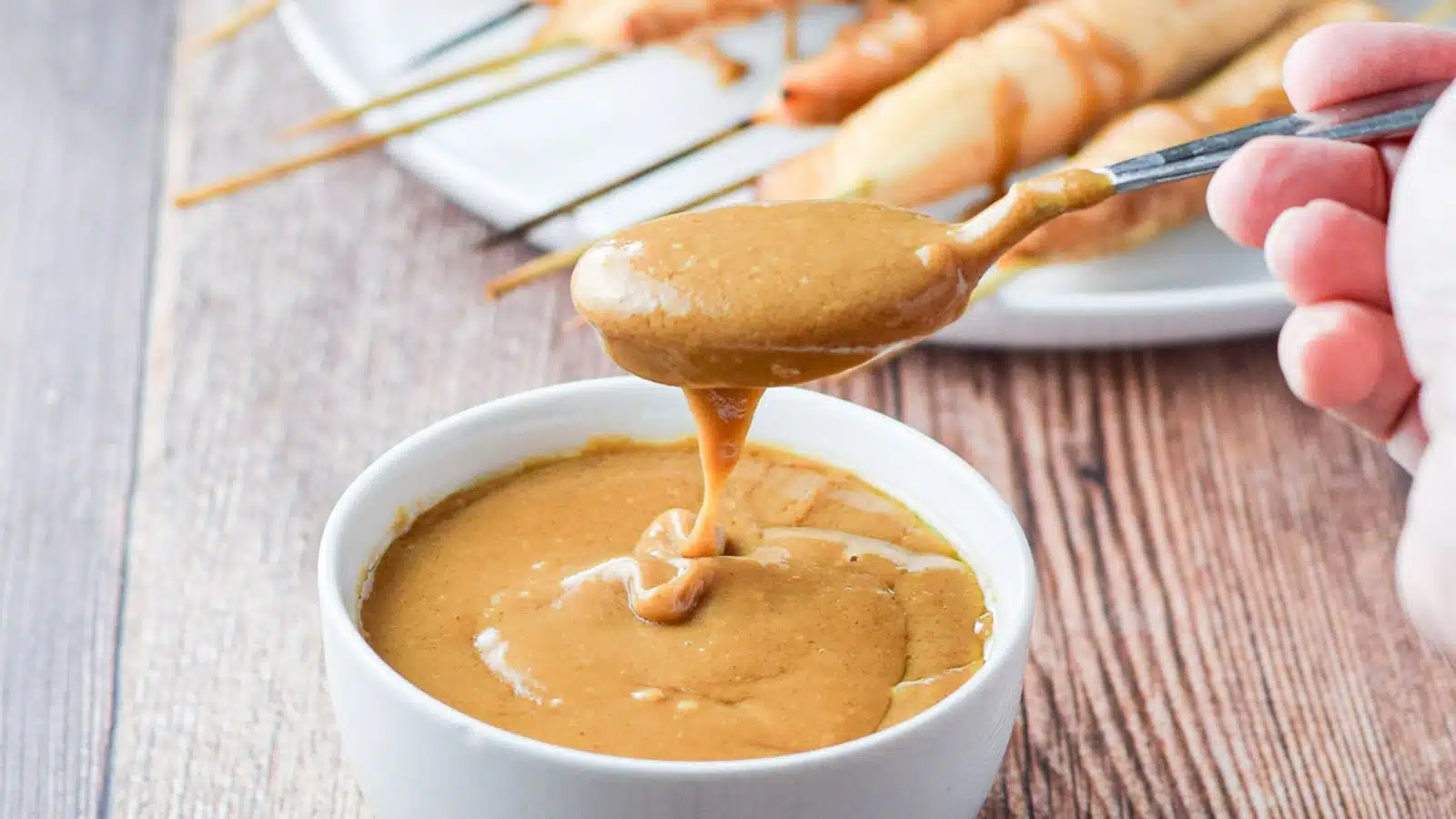 A hand holding a spoon with peanut sauce held over the bowl with chicken behind it