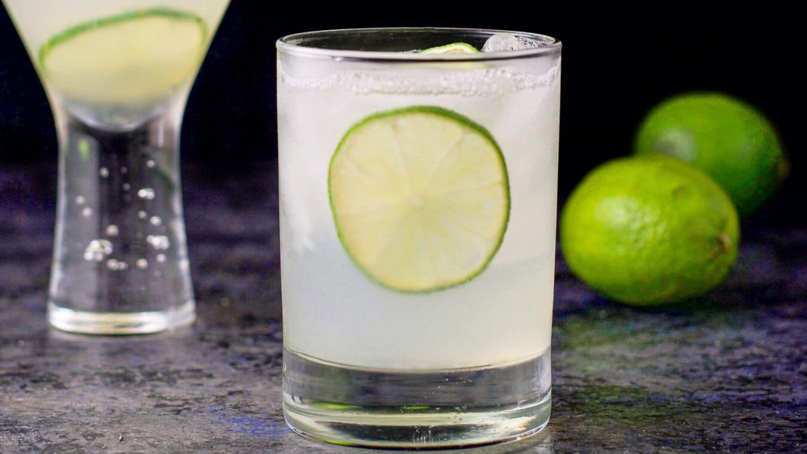 a double old fashioned glass with the gimlet in it along with limes on the table and a lime wheel in the drink