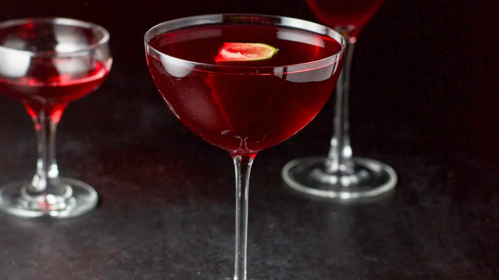 A bowl glass filled with the red cocktail with two others behind with lime wedges in as garnish