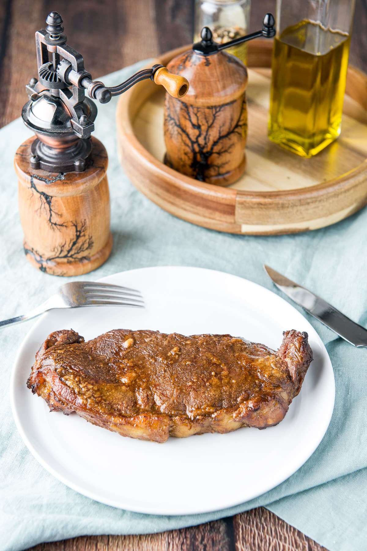 A white plate with steak on it along with a fork and knife with salt and pepper mill on the table