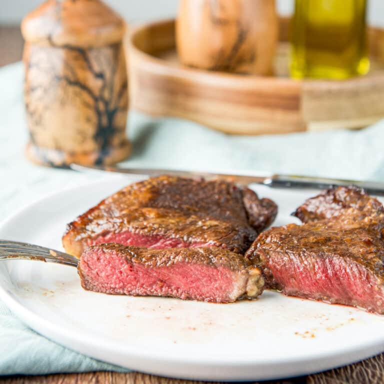 square photo of steak on a white plate cut to show that it's medium rare - with a salt and pepper mill in the background