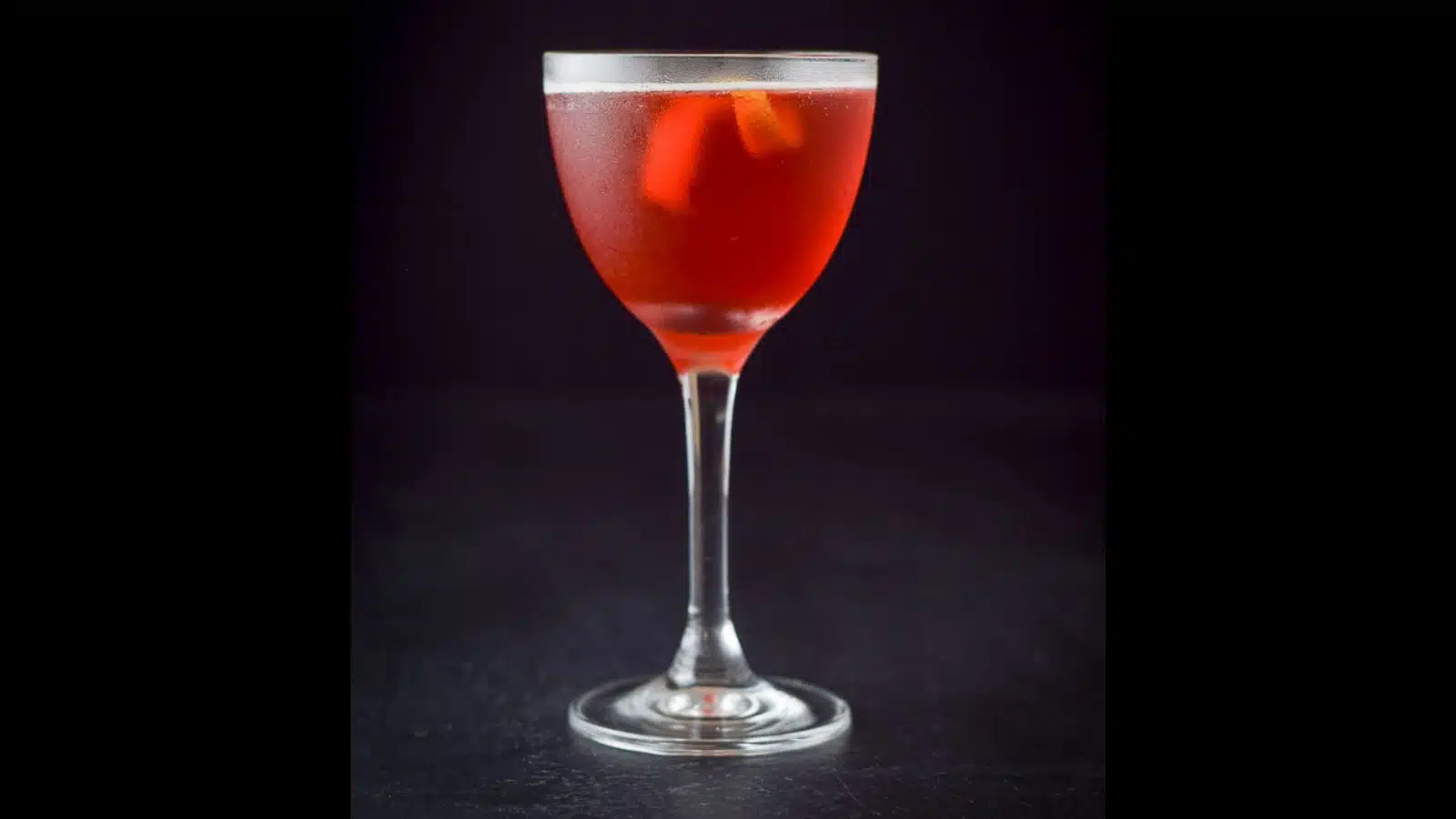 A nick and nora glass filled with the red cocktail with a lemon twist in it
