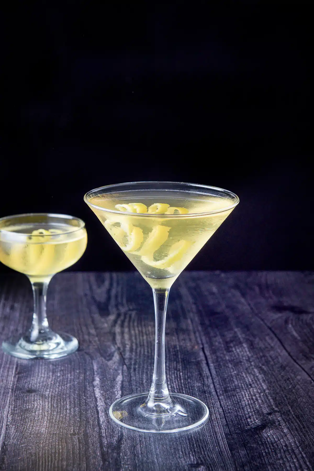 Classic martini glass in front of a coupe glass, filled with the Claridge. They both have lemon twists as garnish