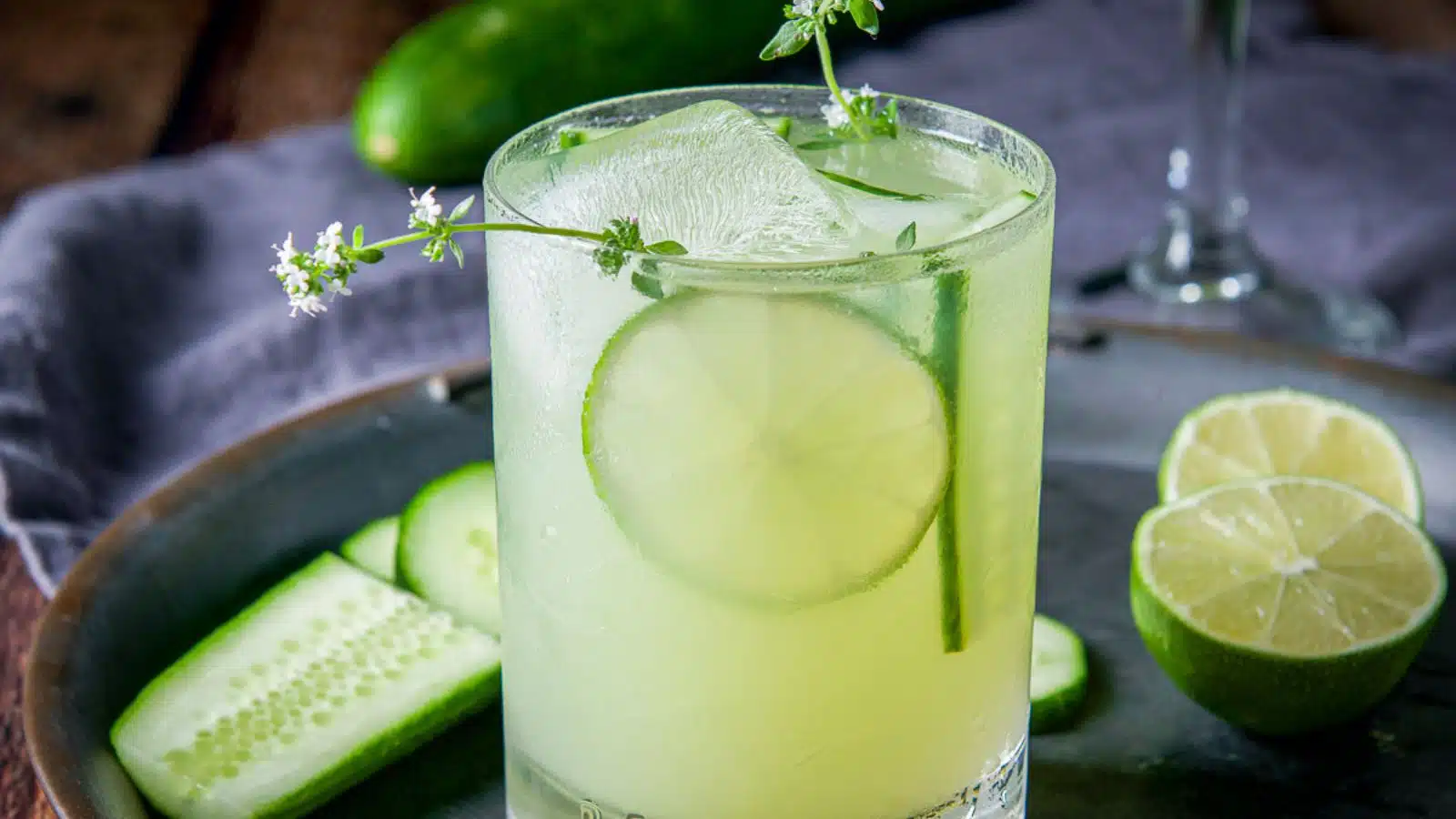 A glass with the margarita in it along with cucumber, an herb and lime wheels