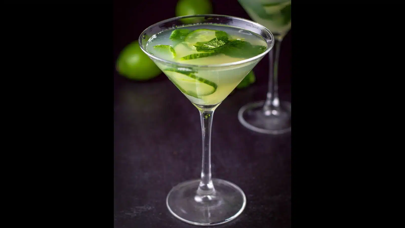 A classic martini glass filled with the cucumber, mint drink with a lime on the table and cucumber and mint in the drink