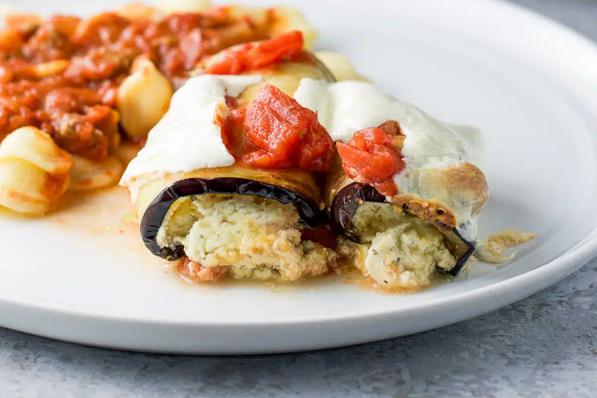 a white plate with stuffed rolled eggplant and pasta with sauce