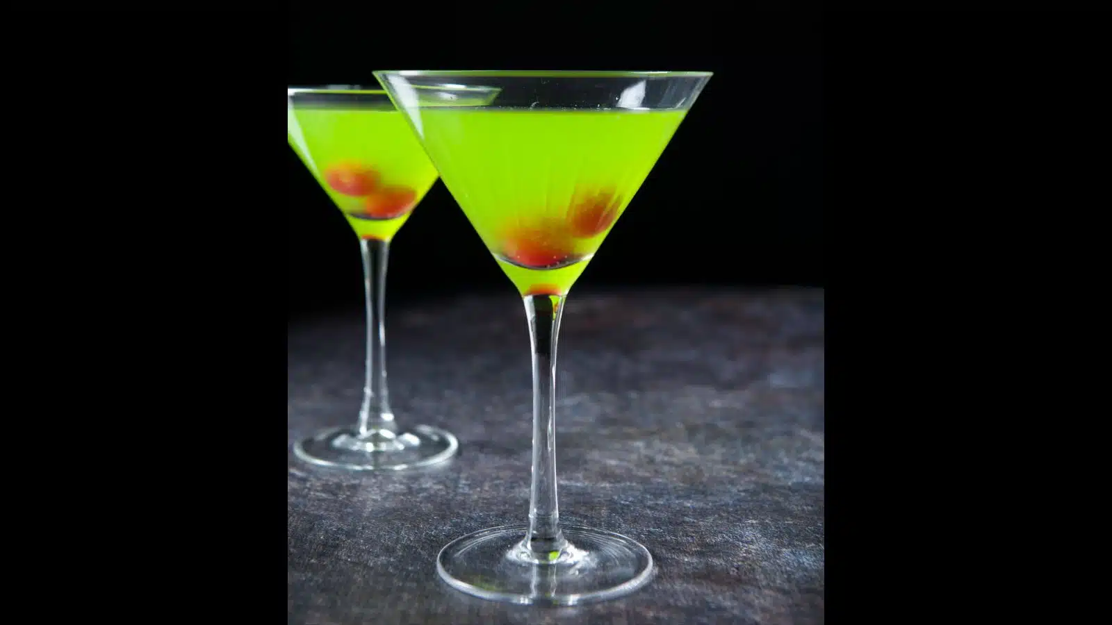 Two martini glasses filled with the bright green cocktail with two cherries in each glass