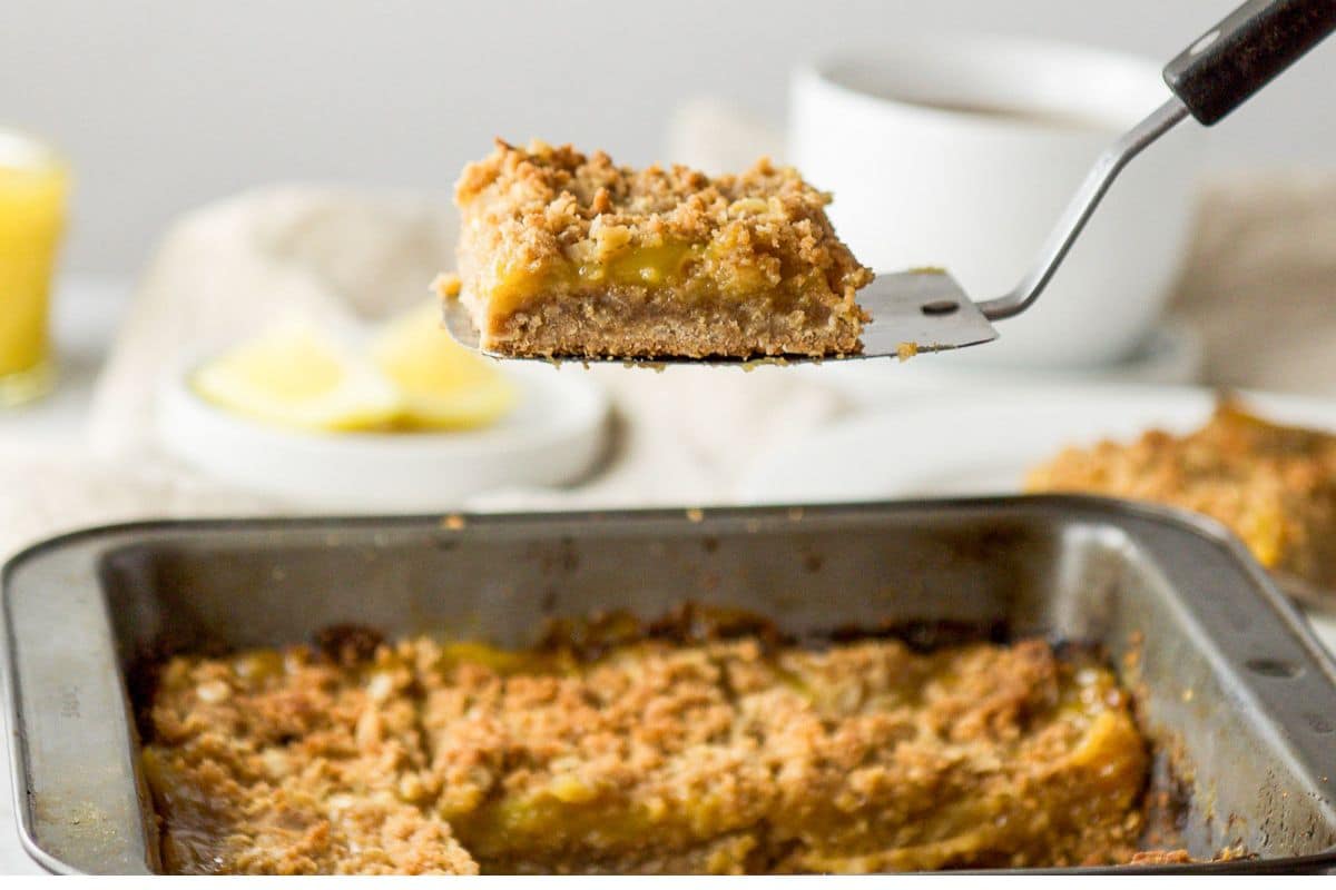 a spatula in the air with a lemon bar on it held over the pan with coffee and lemon in the back