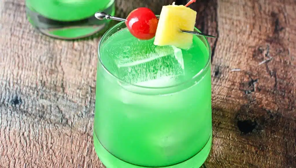 a glass with the green cocktail with a cherry and pineapple chunk as garnish