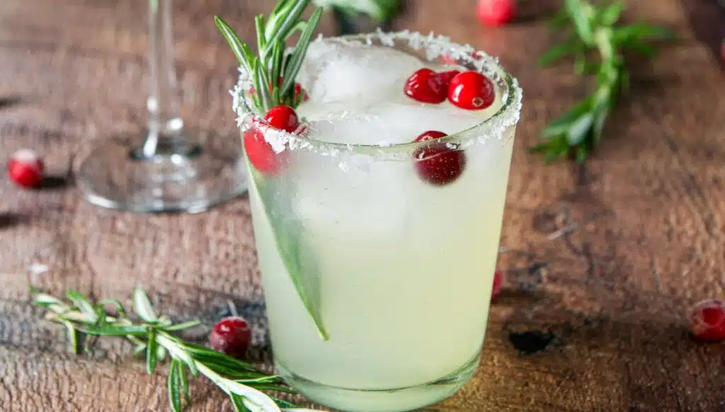 A glass with salt on the rim filled with a margarita with cranberries and rosemary as garnish