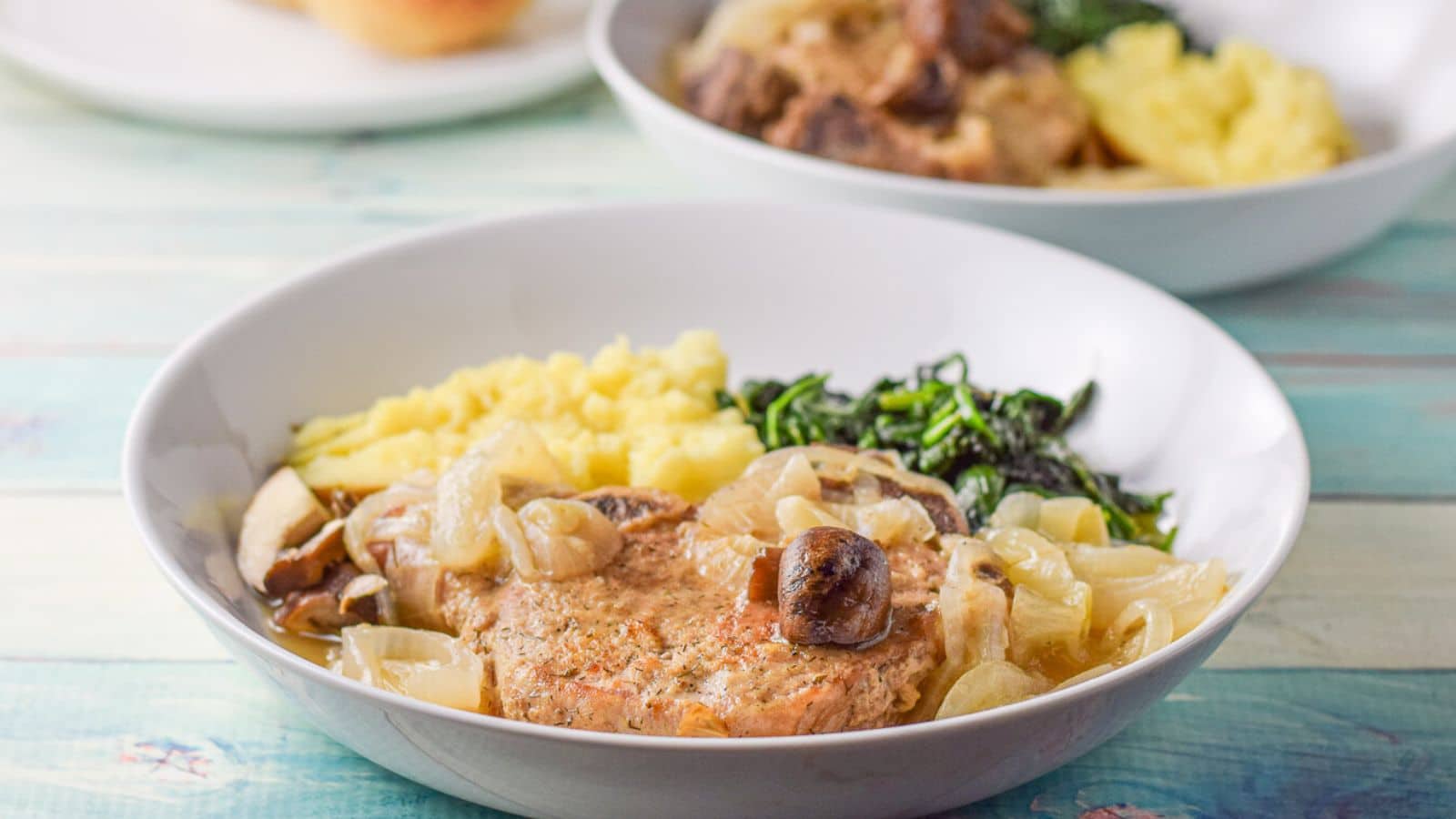 A deep plate with a pork chop with mushrooms, onions and a sauce along with a potato and spinach