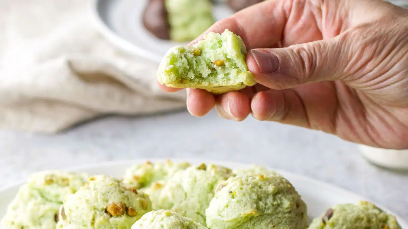 A hand holding a pistachio cookie with a bite out of it held over a plate of cookies