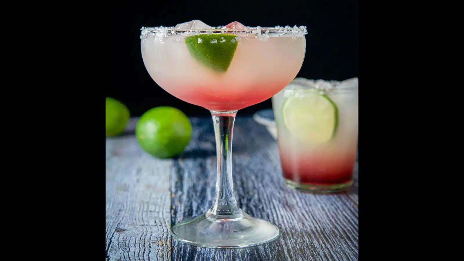 A bowl glass and a double old-fashioned glass filled with a two-toned margarita with limes as garnish