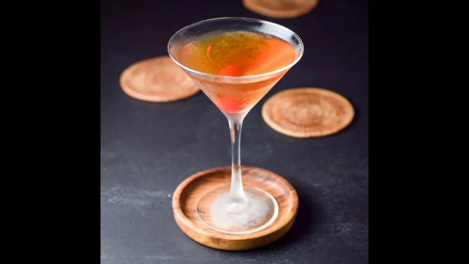 A reddish cocktail in a chilled martini glass with coasters in the back
