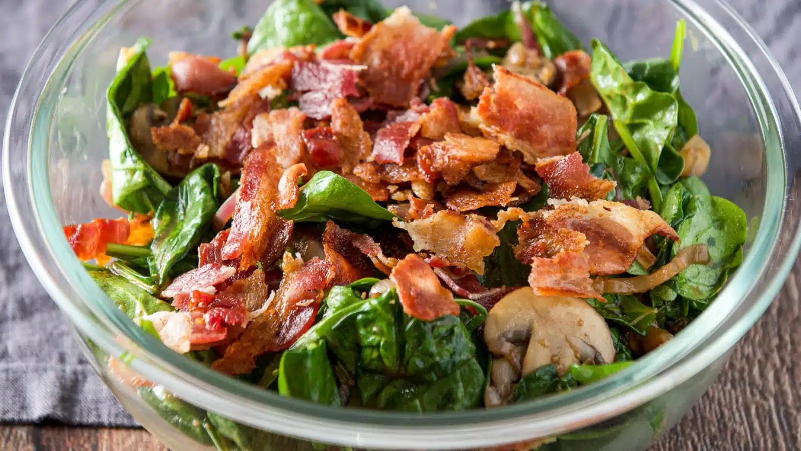 a glass bowl with bacon on a spinach salad