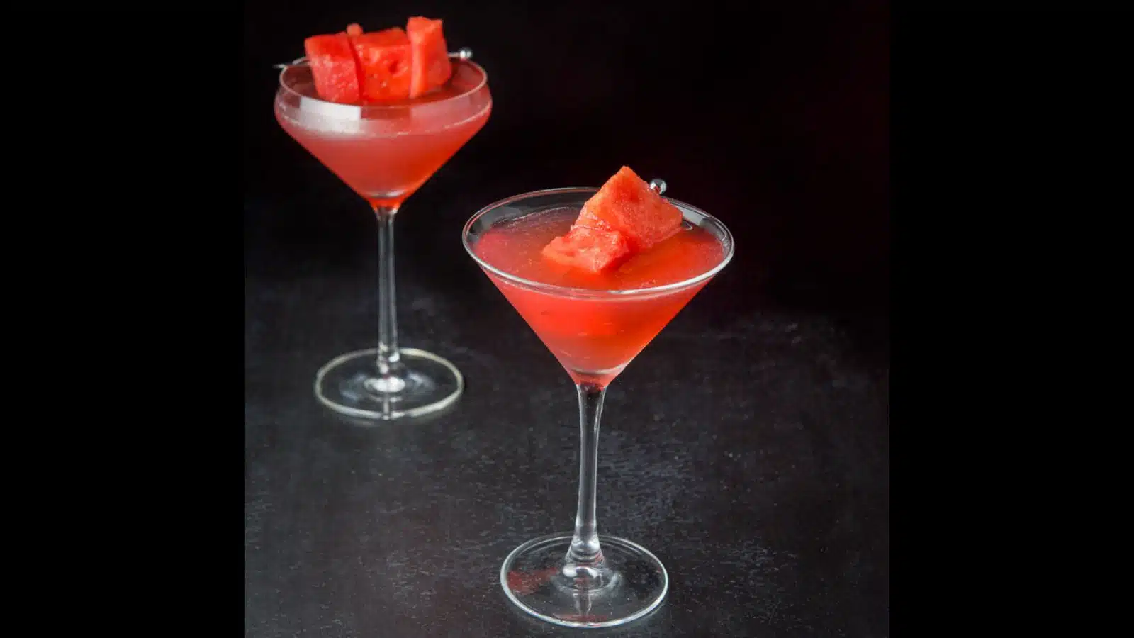 Two martini glasses with the watermelon drink in it along with chunks of watermelon on martini picks