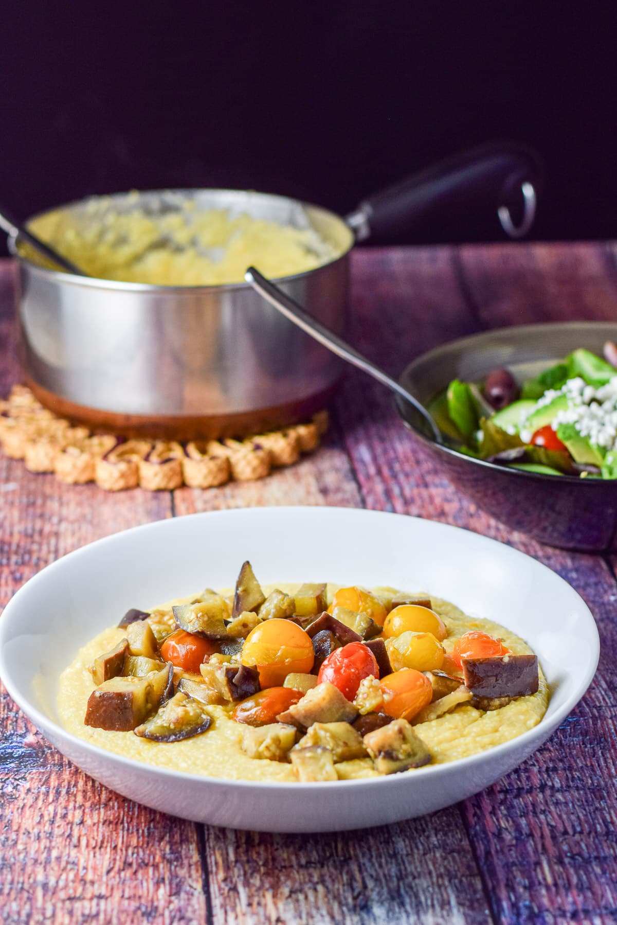 Cooked tomatoes and eggplant on the polenta with a salad and pan of polenta in the background
