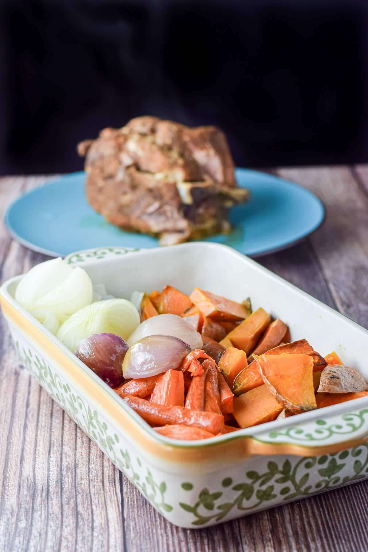 A baking dish filled with cooked vegetables with pulled pork in the back on a blue plate