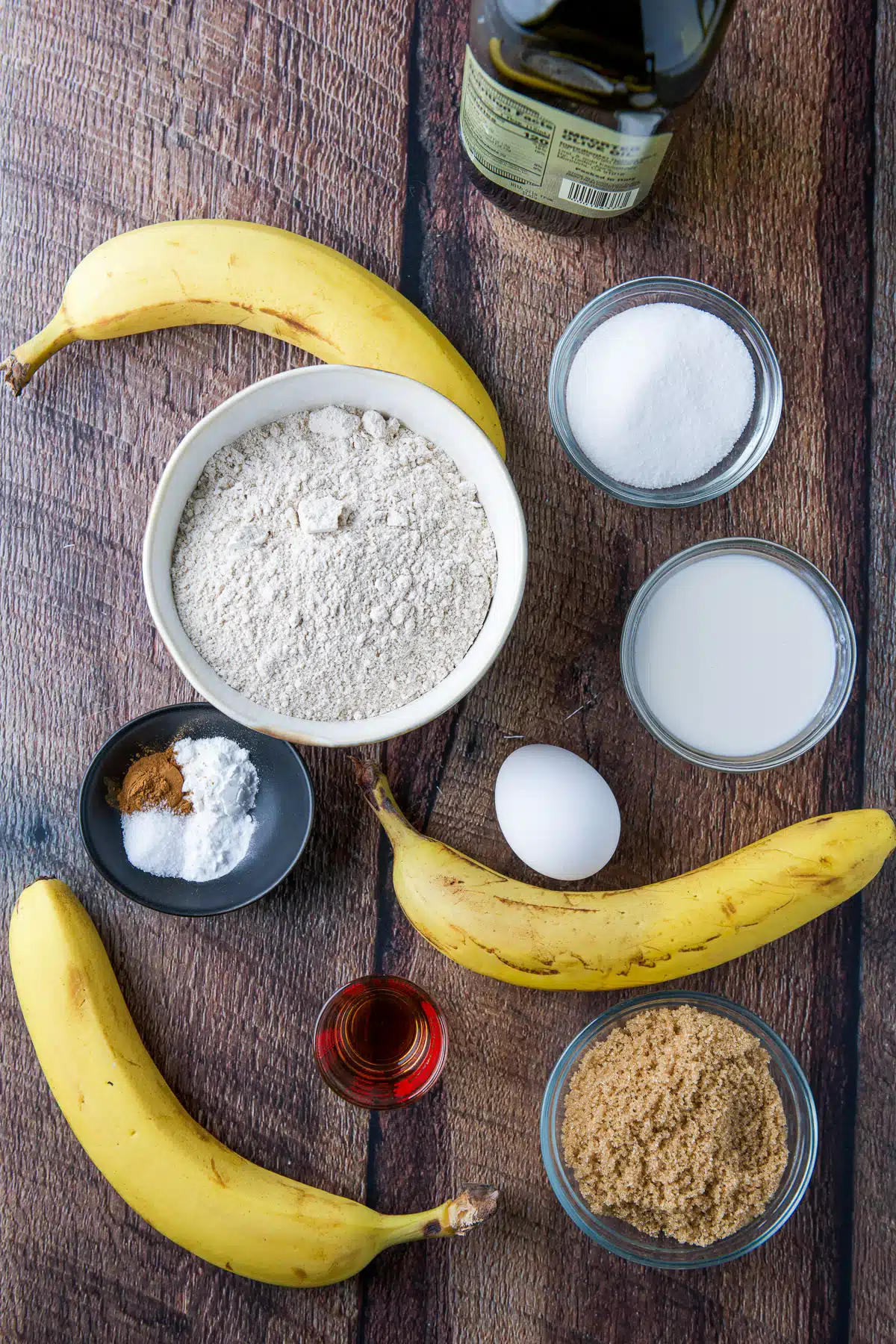 Bananas, sugars, egg, baking powder and soda, cinnamon, oil, milk, and vanilla on a wooden table