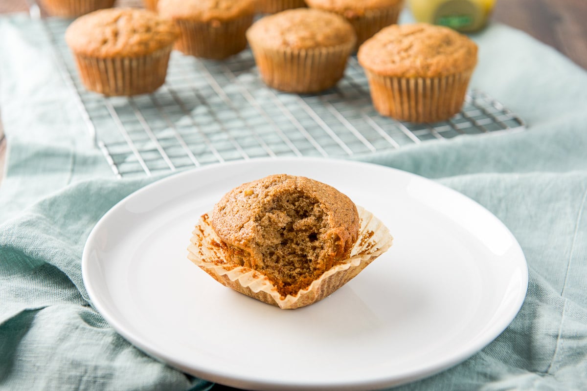 A bite taken out of a muffin still in the wrapper with more muffins in the back on a wire rack