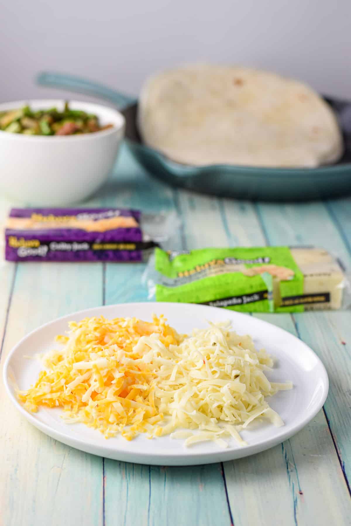 Shredded cheeses on a white plate with cooked vegetable and tortillas in the background