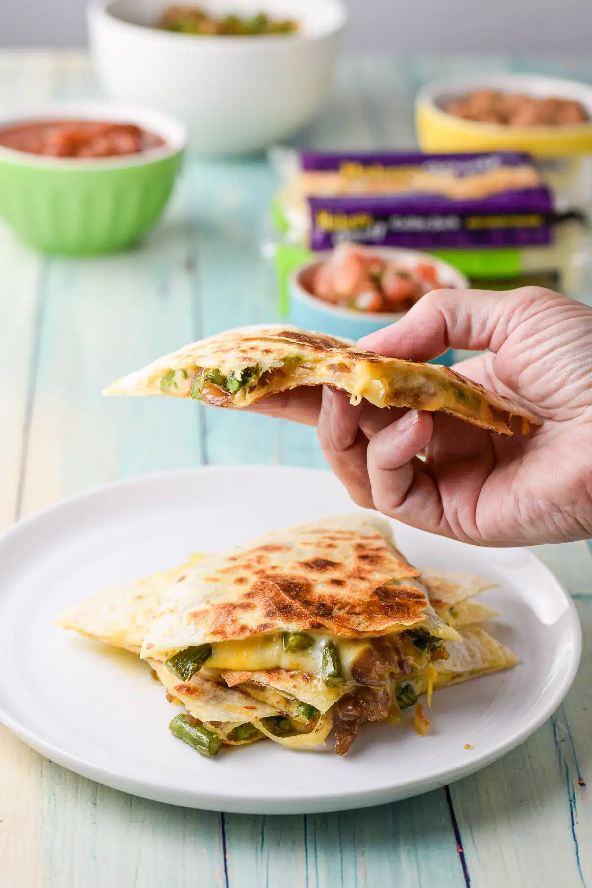 A hand holding a triangle of quesadilla with a bite taken out of it. It is over a plate of more quesadillas