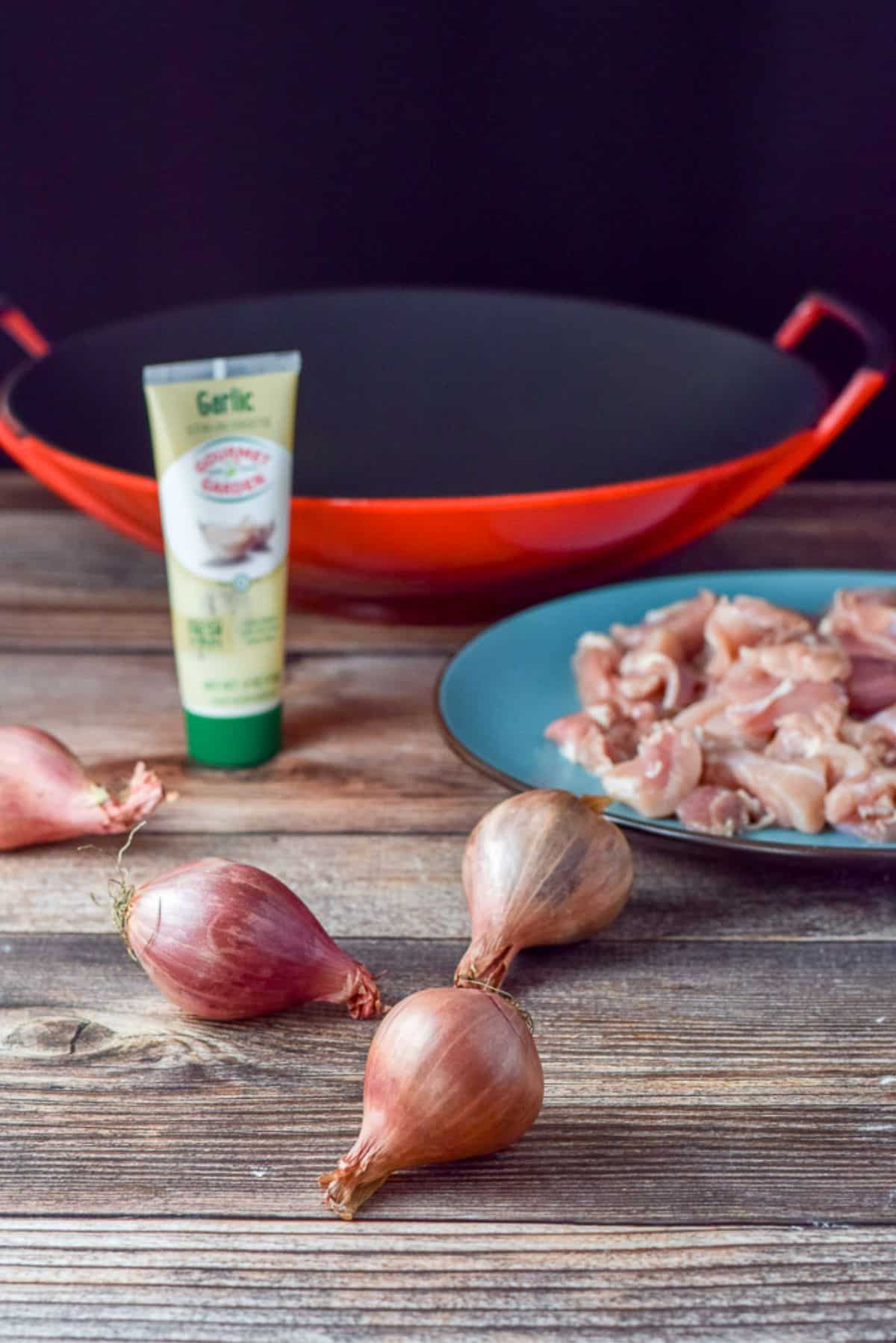 A table with shallots, a plate of raw chicken pieces, garlic, and a wok