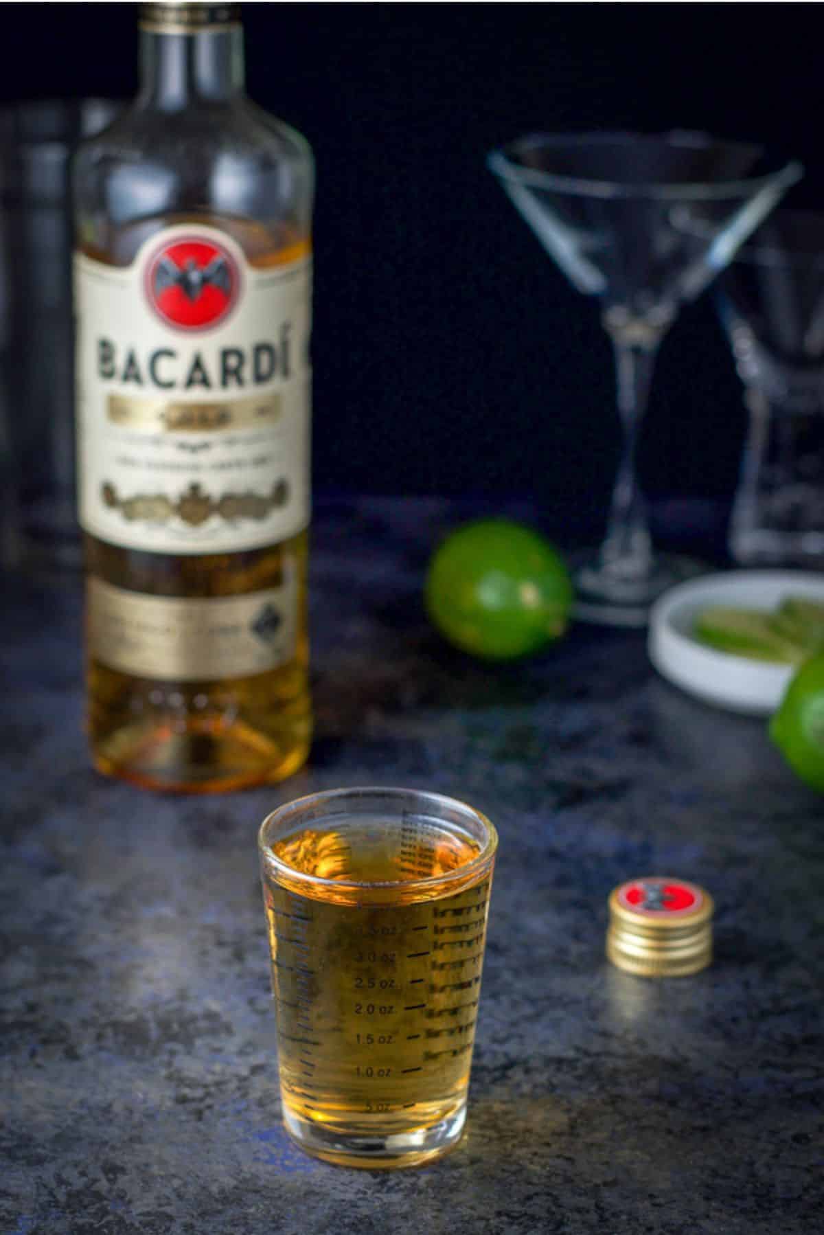 Gold rum measured out with the bottle behind it along with some glasses and lime