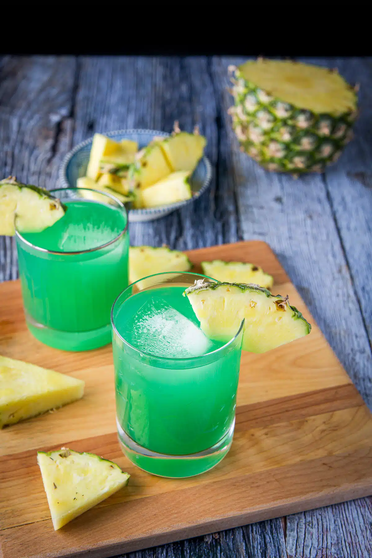 A square wood board with two glasses of a green drink with pineapple wedges as garnish
